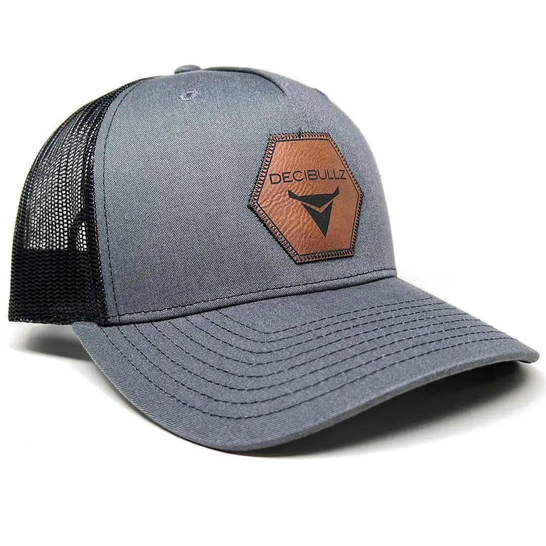 A Decibullz Leather Patch Trucker Hat by Decibullz is a gray and black trucker style hat with a mesh back, featuring a distinctive leather logo patch stitched in a hexagonal shape with the text "DECIBULLZ" and a stylized design. The hat has a curved brim, six-panel construction, and an adjustable snapback for the perfect fit.