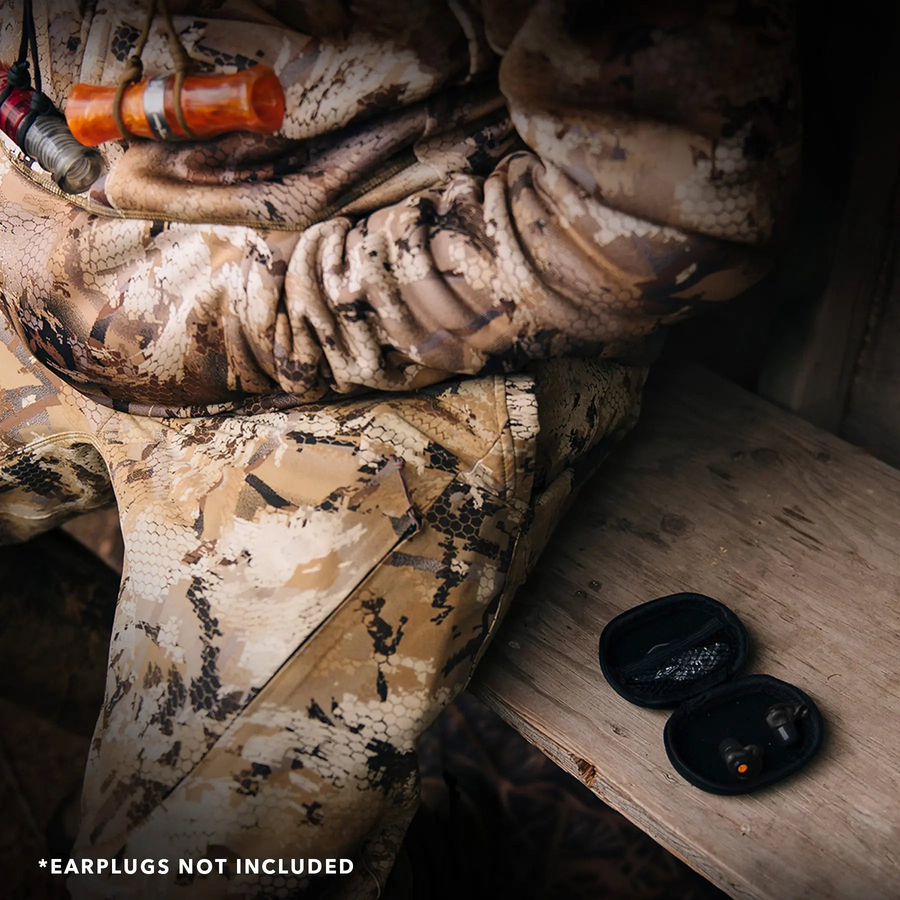 A person in camouflage clothing is sitting on a bench. A small black Decibullz Premium Earplug and Earphone Carrying Case is open and placed beside them on the bench. Note at the bottom of the image states "*EARPLUGS NOT INCLUDED".