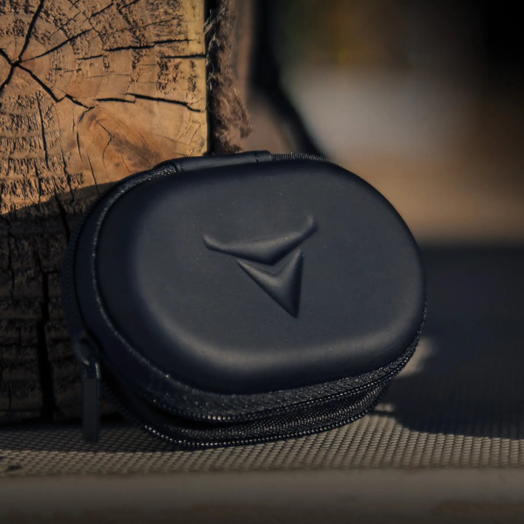 A small black Decibullz Premium Earplug and Earphone Carrying Case with a triangular logo sits against a wooden surface. The case has smooth, rounded edges and appears to be for carrying compact items or accessories like earphones. The background is slightly out of focus, emphasizing the case.
