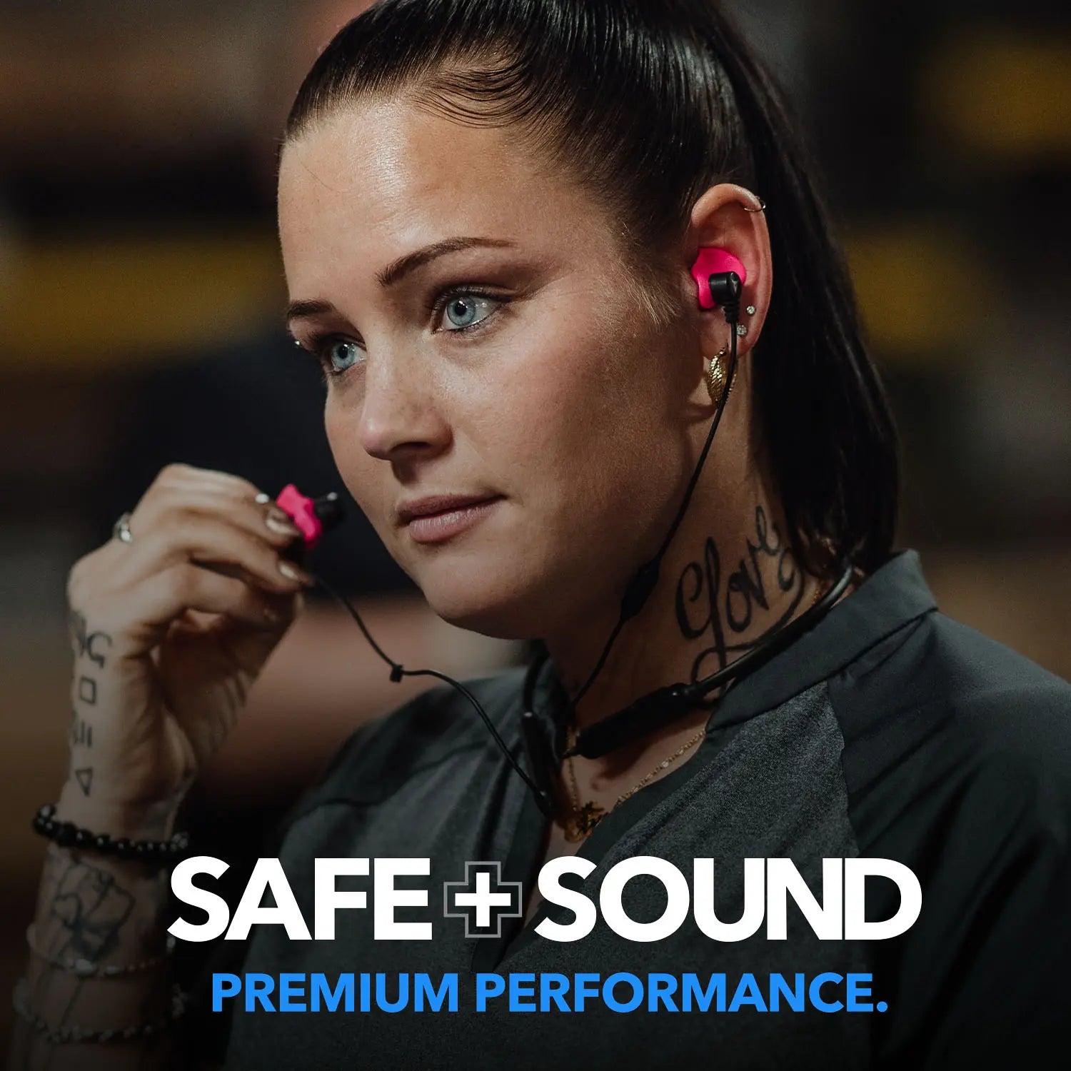 A person with a ponytail is putting pink SAFE + SOUND Custom Molded Bluetooth Wireless Earplug Headphones by Decibullz into their left ear. The text on their neck says "Love," and there is a small tattoo on their wrist. They are wearing a gray top, and the background is blurred. The text reads, "SAFE + SOUND PREMIUM PERFORMANCE.